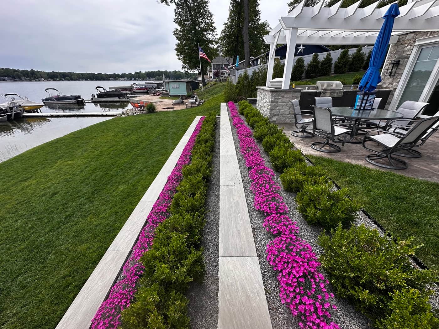 Tiered planting beds with pink impatiens and boxwood along a Southwest Michigan lakefront, lake and pergola in background